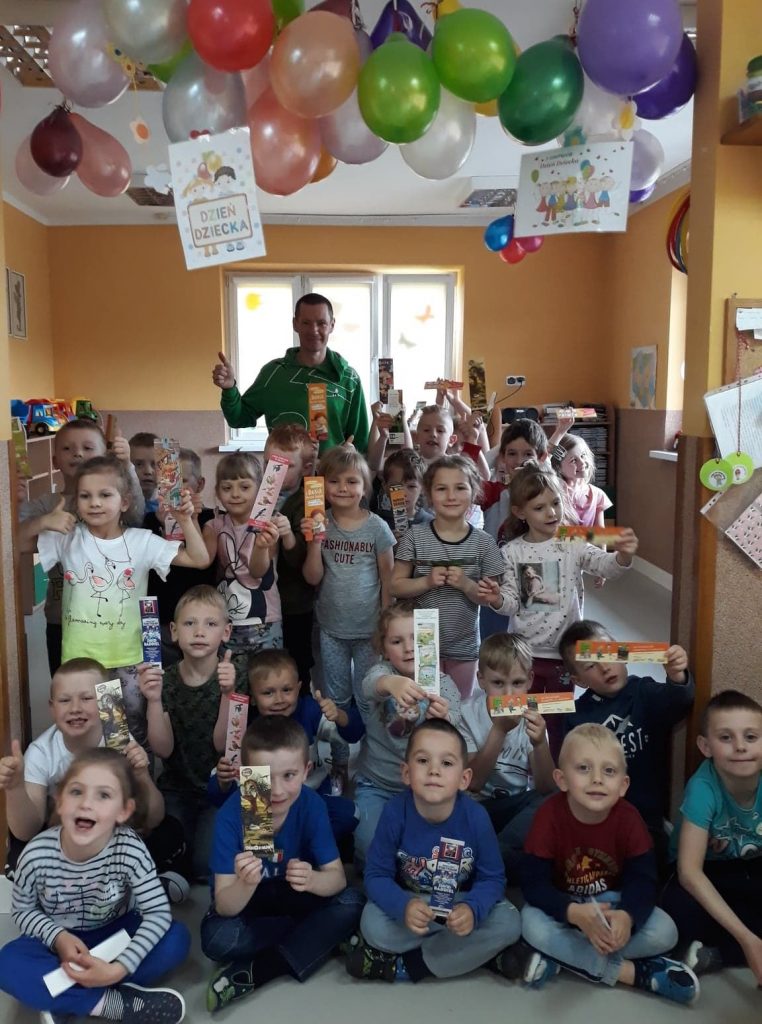 Liczna grupa uśmiechniętych przedszkolaków pozuje razem z bibliotekarzem trzymając w rękach pamiątkowe kolorowe zakładki.