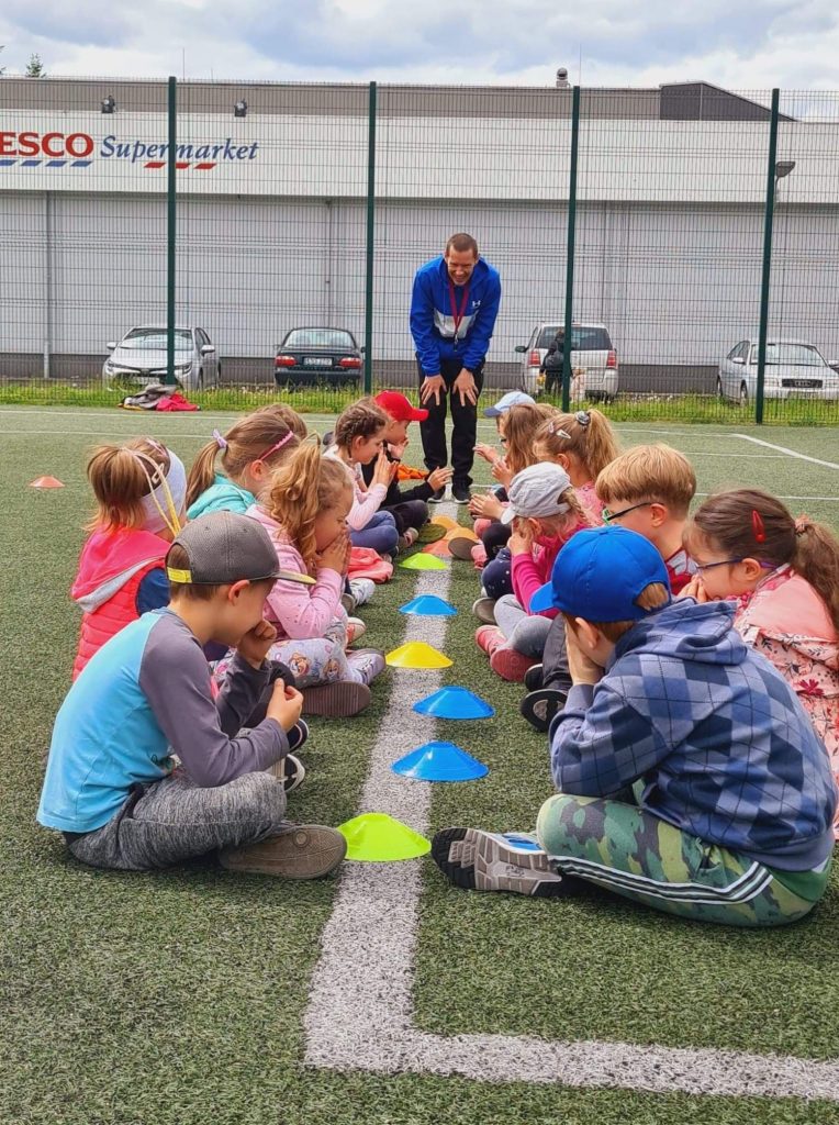 Przedszkolaki dobrane w pary siedzą naprzeciwko siebie na trawie, pomiędzy nimi znajdują się kolorowe plastikowe stożki – „korony”, które trzeba na sygnał włożyć na głowę.