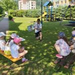Grupa przedszkolaków siedzi wygodnie na ławkach ustawionych na trawniku i słucha z przejęciem bajki o śmiesznym Kaczorku pt. „Mogę być w waszej paczce?”.
