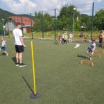 Na znajdującym się obok przedszkola boisku grupa małych sportowców pokonuje ułożony z niskich płotków i słupków sprawnościowy tor przeszkód.