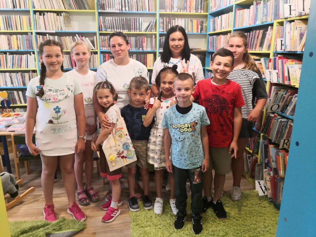 Na fotografii widać grupę osób są to dzieci oraz panie z biblioteki. W tle widać regały z książkami.