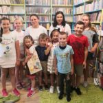 Na fotografii widać grupę osób są to dzieci oraz panie z biblioteki. W tle widać regały z książkami.