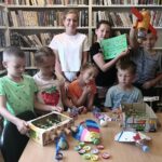 Na fotografii widać grupkę dzieci, dziewczynki i chłopcy stojących na tle regału z książkami. Przed nimi widoczny jest stół na którym widoczne są różnego rodzaju przedmioty- są to zabawki wykonane przez dzieci ze śmieci np. plastikowych butelek czy kartonowych pudełek