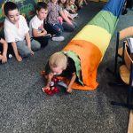 Na zdjęciu widać kolorową salę przedszkolną, dzieci siedzą na wykładzinie. Na ziemi rozwinięty jest tunel z kolorowej tkaniny. Wdać dziewczynkę która przeciska się przez ten tunel. Pozostałe dzieci obserwują z uśmiechami zabawę.
