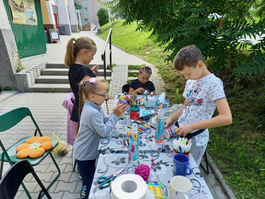 Za zewnatrz biblioteki przy długim stole stoją trzy dziewczynki i jeden chłopiec.