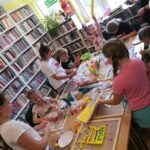 Na zdjęciu widać dzieci stojące przy stole w czytelni biblioteki. Każdy zajęty jest tworzeniem ścieżki sensorycznej z kolorowych patyczków, koralików papieru, kamyków, piórek