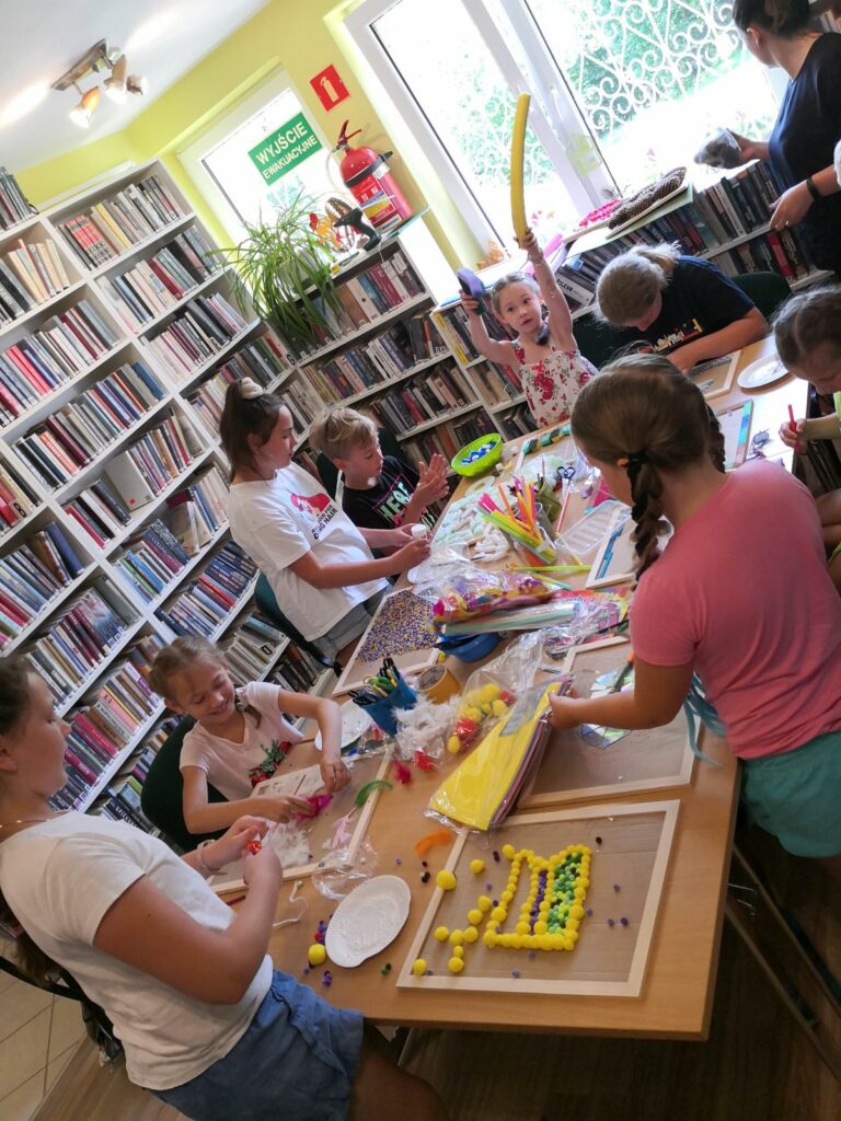Na zdjęciu widać dzieci stojące przy stole w czytelni biblioteki. Każdy zajęty jest tworzeniem ścieżki sensorycznej z kolorowych patyczków, koralików papieru, kamyków, piórek