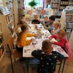 Dziewczynki i chłopcy siedzą przy długim stole i wybierają kamienie, które za chwilę pięknie pomalują farbami.