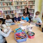 Na zdjęciu widać grupę dzieci siedzących przy stole w sali bibliotecznej. Dookoła widać regały z książkami oraz czytająca bibliotekarkę.