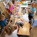 Dzieci siedzą przy długim stole i przy pomocy szyszek, nakrętek, kamyczków, folii, czy kasztanów komponują ścieżkę sensoryczną.