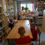 W czytelni biblioteki dzieci siedzą przy stole i słuchają jak bibliotekarz czyta im jeden z rozdziałów Eko- książki „Śmieciogród”.