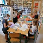 Grupa uczestników zajęć pracuje przy stole nad swoimi zabawkowymi samolotami. Dzieci wykorzystują m.in. plastikowe butelki, kolorowy papier, nakrętki i tekturę.