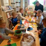 Praca przy stole idzie pełną parą. Grupa uczestników wakacyjnych warsztatów razem z bibliotekarzem kleją, wycinają i kolorują plastikowe doniczki.