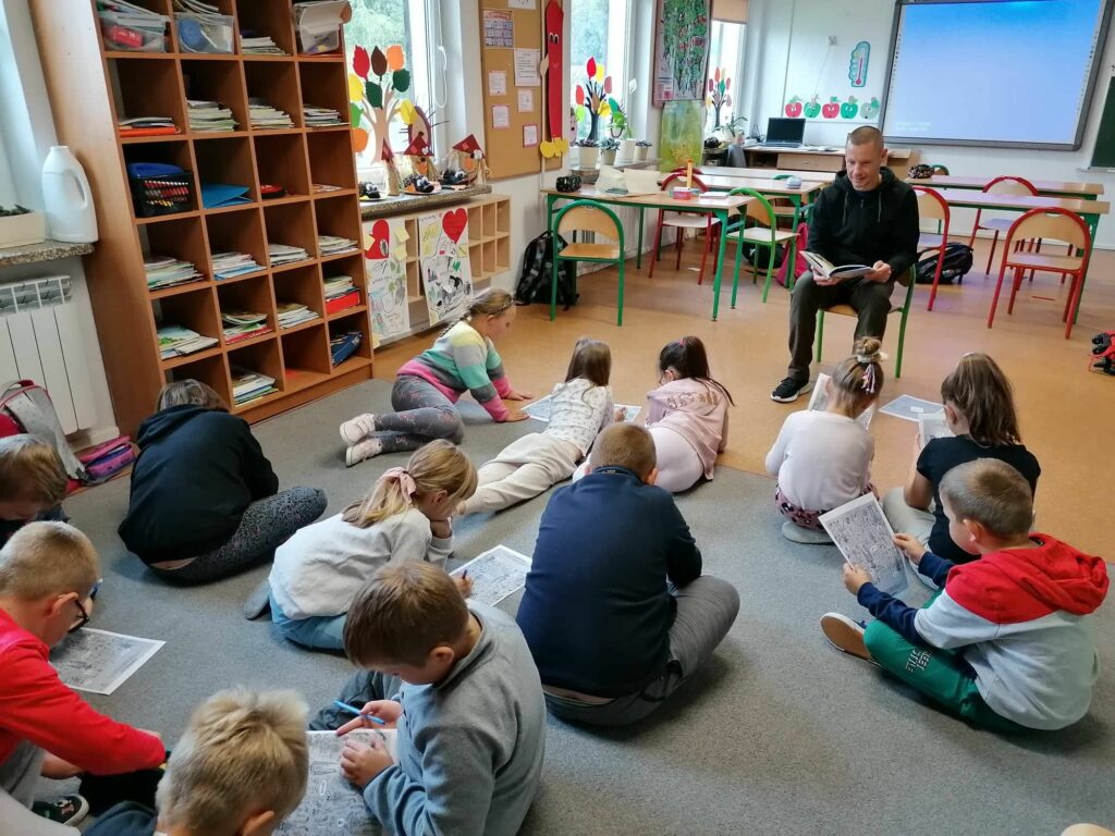 W jednej z klas uczniowie siedzą na dywanie, rozwiązują rebusy na kartkach i słuchają pana bibliotekarza udzielającego im wskazówek z książki.