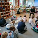 W jednej z klas uczniowie siedzą na dywanie, rozwiązują rebusy na kartkach i słuchają pana bibliotekarza udzielającego im wskazówek z książki.