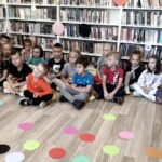 Na zdjęciu widać dzieci siedzące na podłodze i słuchające czytanki. Przed dziećmi na podłodze widoczne są kolorowe kropki wycięte z papieru. za nimi widać regały z książkami.
