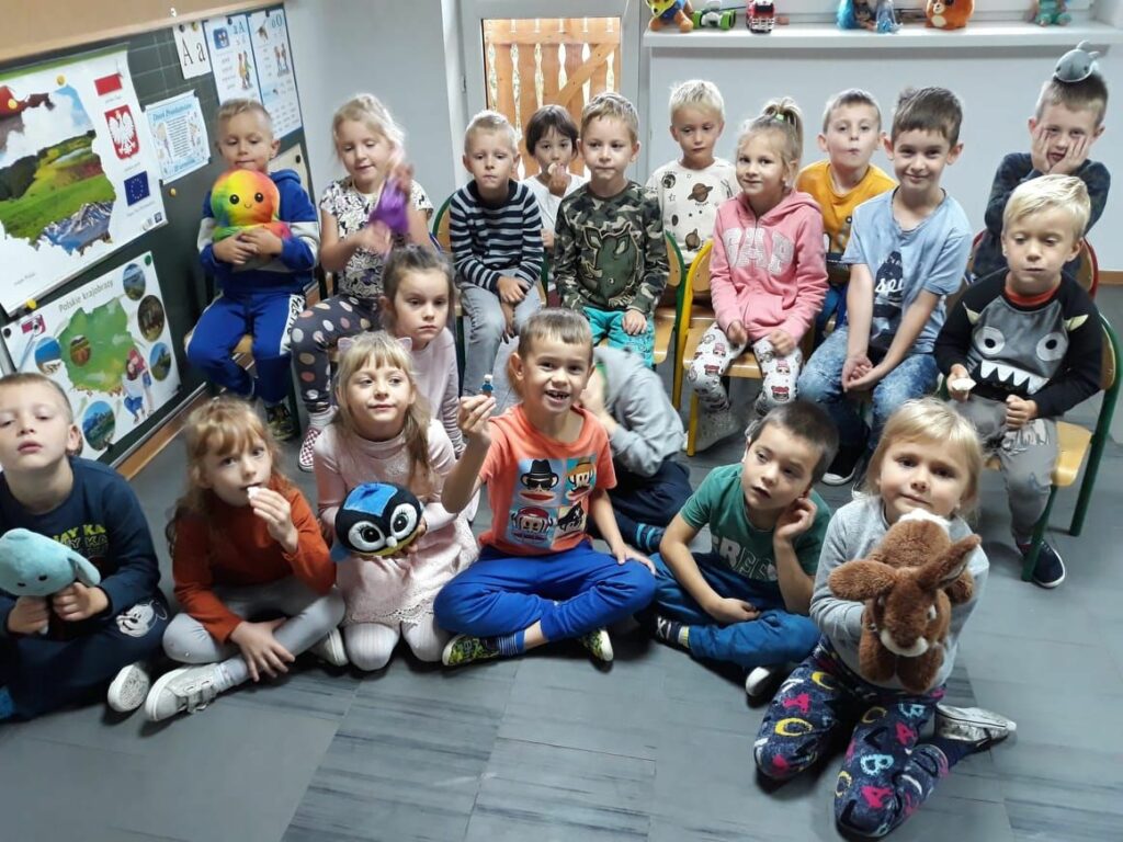 Grupa przedszkolaków siedzi grzecznie w sali i trzymając w rękach swoje ulubione zabawki słucha z uwagą książeczki pt. „Posłuchaj, mały!”