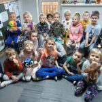 Grupa przedszkolaków siedzi grzecznie w sali i trzymając w rękach swoje ulubione zabawki słucha z uwagą książeczki pt. „Posłuchaj, mały!”