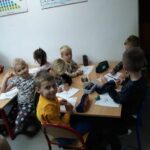 Dzieci siedzą przy malutkich stolikach i przy pomocy kredek kolorują swoje rysunki.