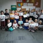 Uśmiechnięte przedszkolaki ustawiły się pod jedną ze ścian do pamiątkowego zdjęcia. Wszyscy trzymają w rękach wykonane na zajęciach piękne rysunki.