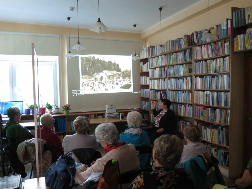 Pani dyrektor biblioteki przedstawia wyświetlane kolejne ilustracje kuracjuszom siedzącym w sali czytelni