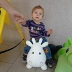 Na nadmuchanej krówce zabawce siedzi mała dziewczynka okoł jednego roku. Widac ręke podtrzymującą dziecko.