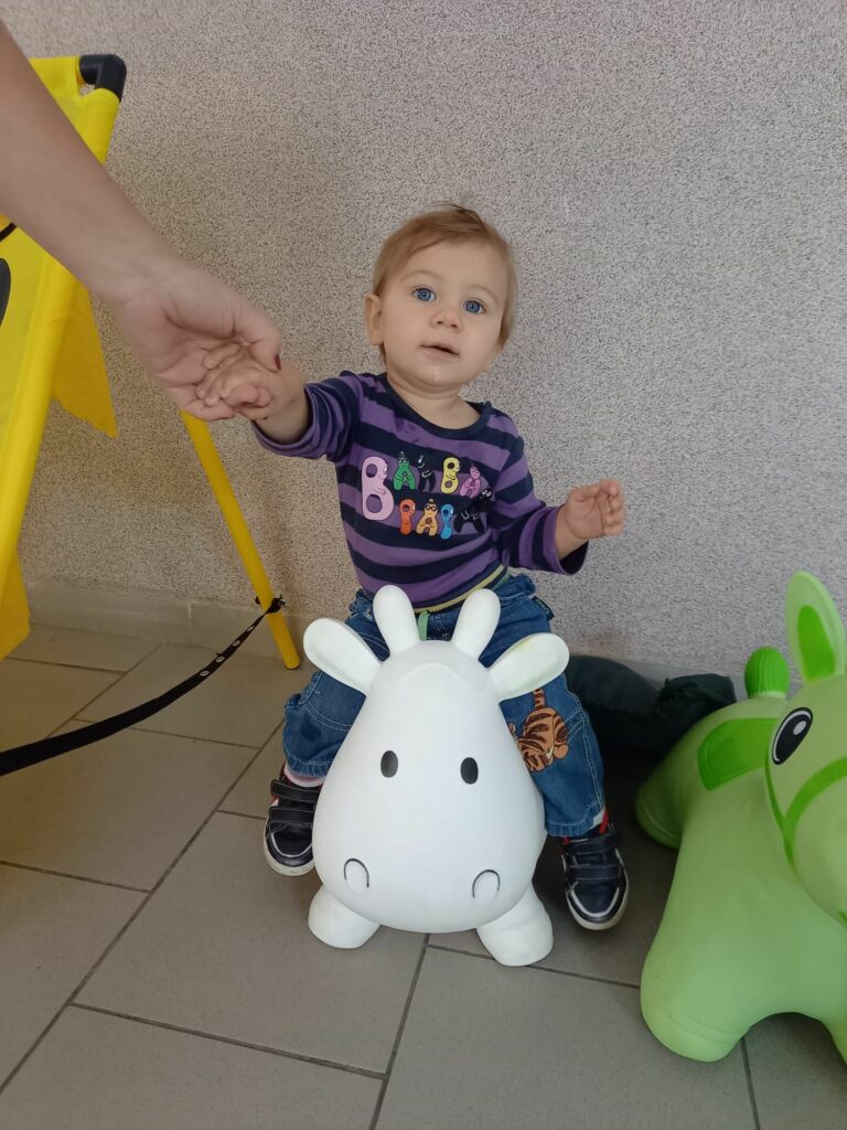 Na nadmuchanej krówce zabawce siedzi mała dziewczynka okoł jednego roku. Widac ręke podtrzymującą dziecko.