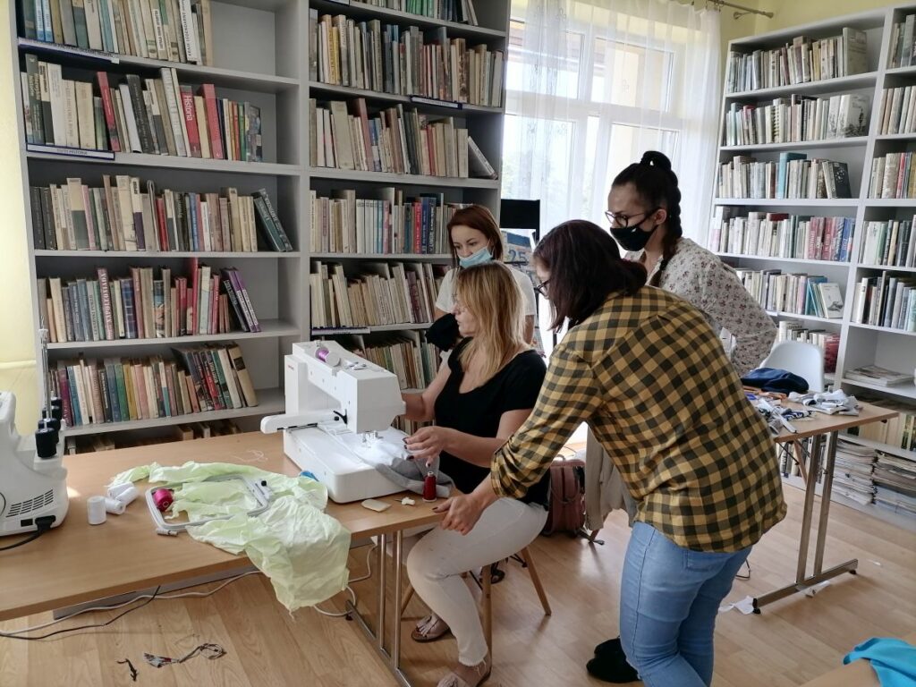 Na zdjęciu pomieszczenie biblioteki. Wokół na ścianach regały z książkami. Przy stole siedzi kobieta o obsługuje maszynę do szycia hafciarkę. W okół niej stoją trzy młode kobiety i przyglądaja się.