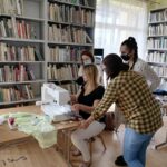 Na zdjęciu pomieszczenie biblioteki. Wokół na ścianach regały z książkami. Przy stole siedzi kobieta o obsługuje maszynę do szycia hafciarkę. W okół niej stoją trzy młode kobiety i przyglądaja się.