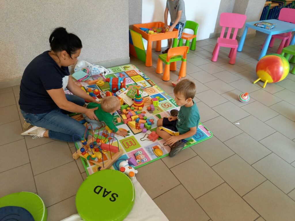 Na kolorowej macie siedzi jedna z pań bibliotekarek w średnim wieku i opiekuje sie dwoma małymi chłopcami. Na macie rozłożone