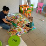 Na kolorowej macie siedzi jedna z pań bibliotekarek w średnim wieku i opiekuje sie dwoma małymi chłopcami. Na macie rozłożone