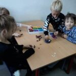 Trzy dziewczynki i jeden chłopczyk siedzą przy malutkim stoliczku na którym znajduje się pudełko z plasteliną i kasztany z których dzieci robią piękne zwierzątka.