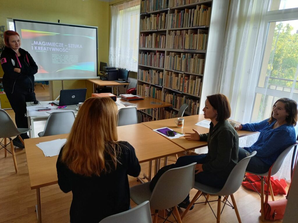 Na zdjęciu widać 3 uczestniczki projektu "MAMY-to!" oraz Panią prowadzącą zajęcia z doradztwa zawodowego. Panie siedzą przy stolikach, twarzami skierowanymi w stronę ekranu, na którym wyświetlana jest prezentacja. Na stolikach widoczne są kartki papieru, długopisy, projektor oraz laptop. Zajęcia odbywają się w bibliotece - w tle widoczne są regały z książkami.