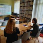 Na zdjęciu widać 3 uczestniczki projektu "MAMY-to!" oraz Panią prowadzącą zajęcia z doradztwa zawodowego. Panie siedzą przy stolikach, twarzami skierowanymi w stronę ekranu, na którym wyświetlana jest prezentacja. Na stolikach widoczne są kartki papieru, długopisy, projektor oraz laptop. Zajęcia odbywają się w bibliotece - w tle widoczne są regały z książkami.