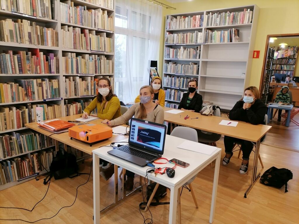 Na zdjęciu widać 5 uczestniczek projektu "MAMY-to!" podczas warsztatów z doradcą zawodowym. Panie siedzą przy stolikach, na których widoczne są kartki papieru, długopisy, projektor oraz laptop. Zajęcia odbywają się w bibliotece - w tle widoczne są regały z książkami.