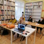 Na zdjęciu widać 5 uczestniczek projektu "MAMY-to!" podczas warsztatów z doradcą zawodowym. Panie siedzą przy stolikach, na których widoczne są kartki papieru, długopisy, projektor oraz laptop. Zajęcia odbywają się w bibliotece - w tle widoczne są regały z książkami.
