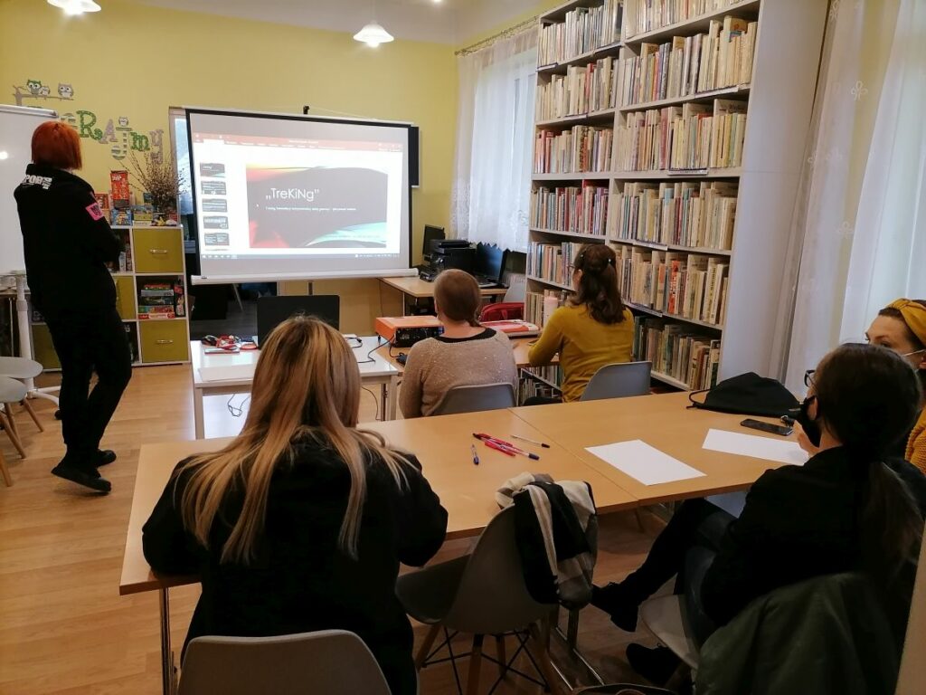 Na zdjęciu widać 5 uczestniczek projektu "MAMY-to!" oraz panią prowadzącą warsztaty z doradztwa zawodowego. Panie siedzą przy stolikach, twarzami skierowanymi w stronę ekranu projekcyjnego, na którym wyświetlana jest prezentacja. Na stolikach widoczne są kartki papieru, długopisy, projektor oraz laptop. Zajęcia odbywają się w bibliotece - w tle widoczne są regały z książkami oraz grami planszowymi.