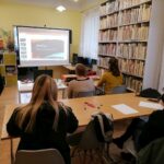 Na zdjęciu widać 5 uczestniczek projektu "MAMY-to!" oraz panią prowadzącą warsztaty z doradztwa zawodowego. Panie siedzą przy stolikach, twarzami skierowanymi w stronę ekranu projekcyjnego, na którym wyświetlana jest prezentacja. Na stolikach widoczne są kartki papieru, długopisy, projektor oraz laptop. Zajęcia odbywają się w bibliotece - w tle widoczne są regały z książkami oraz grami planszowymi.