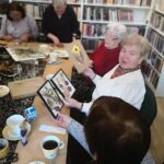 Na zdjęciu widać biblioteczną sale, regału z książkami oraz stoły przy których siedzą seniorki wykonujące prace - obrazki z suszonych roślin.