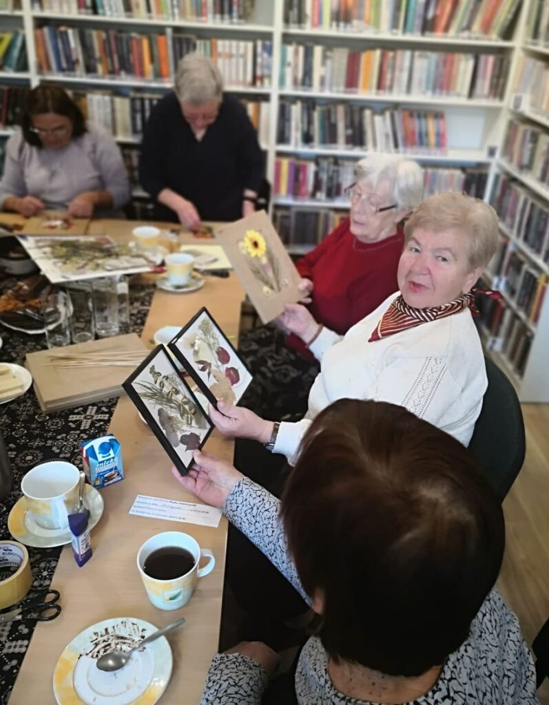Na zdjęciu widać biblioteczną sale, regału z książkami oraz stoły przy których siedzą seniorki wykonujące prace - obrazki z suszonych roślin.