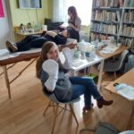 Na zdjęciu widać dwie uczestniczki projektu "MAMY-to!" oraz panią kosmetyczkę. Jedna z uczestniczek leży na specjalnie przygotowanym łóżku, a pani kosmetyczka nakłada jej maseczkę na twarz. Z kolei druga z uczestniczek prezentuje dłonie w specjalnych rękawicach służących do wykonania zabiegu parafinowego na dłoniach. Dodatkowo na stolikach widoczne są kosmetyki używane przez panią kosmetyczkę. Zajęcia odbywają się w bibliotece, w tle widoczne są regały z książkami, wiszący na ścianie telewizor oraz stolik kawowy z drobnym poczęstunkiem.