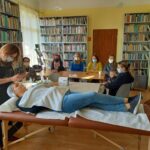 Na zdjęciu widać 7 uczestniczek projekty "MAMY-to!", jedno dziecko - chłopca siedzącego na kolanach mamy oraz panią kosmetyczkę przeprowadzającą zabieg pielęgnacji twarzy u jednej z uczestniczek projektu. Sześć pań siedzi przy stolikach i obserwuje panią kosmetyczkę podczas nakładania maseczki na twarz jednej z koleżanek, która leży na specjalnie przygotowanym łóżku. Spotkanie odbywa się w bibliotece, w tle widoczne są regały z książkami.