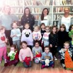 Na zdjęciu widać salę biblioteczną oraz dzieci w wieku przedszkolnym. Kilkoro dzieci wraz z paniami stoi, przed nimi inne dzieci siedzą trzymając w rekach kolorowe pluszaki przedstawiające owoce i warzywa.