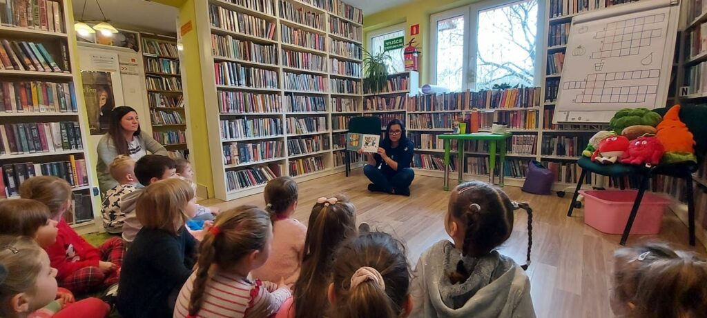 Na zdjęciu widać dzieci siedzące na podłodze w sali bibliotecznej, dzieci słuchają książki czytanej przez bibliotekarkę. Na fotografii widać również regały z książkami, oraz pluszowe owoce.