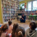 Na zdjęciu widać dzieci siedzące na podłodze w sali bibliotecznej, dzieci słuchają książki czytanej przez bibliotekarkę. Na fotografii widać również regały z książkami, oraz pluszowe owoce.