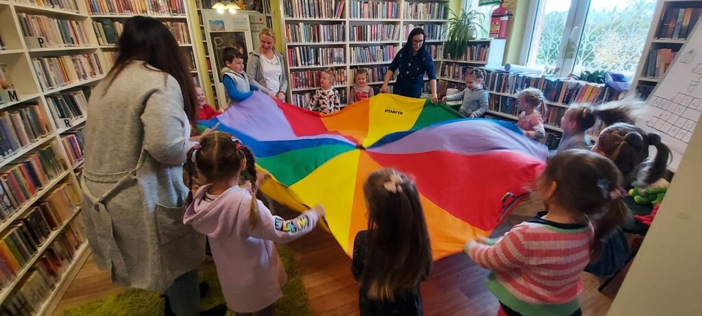 Na zdjęciu widać salę biblioteczną oraz dzieci w wieku przedszkolnym bawiące się kolorową chusta w kształcie koła. Z dziećmi bawią się również trzy panie.