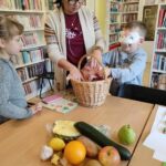 na zdjęciu widać dwoje dzieci dzieci: dziewczynkę i chłopca, widać również bibliotekarkę. Chłopiec ma zasłonięte oczy i próbuje odgadnąć jaki owoc trzyma w rękach. Na stole widać kosz oraz różnego rodzaju owoce i warzywa np. banan, jabłko czy cukinia.