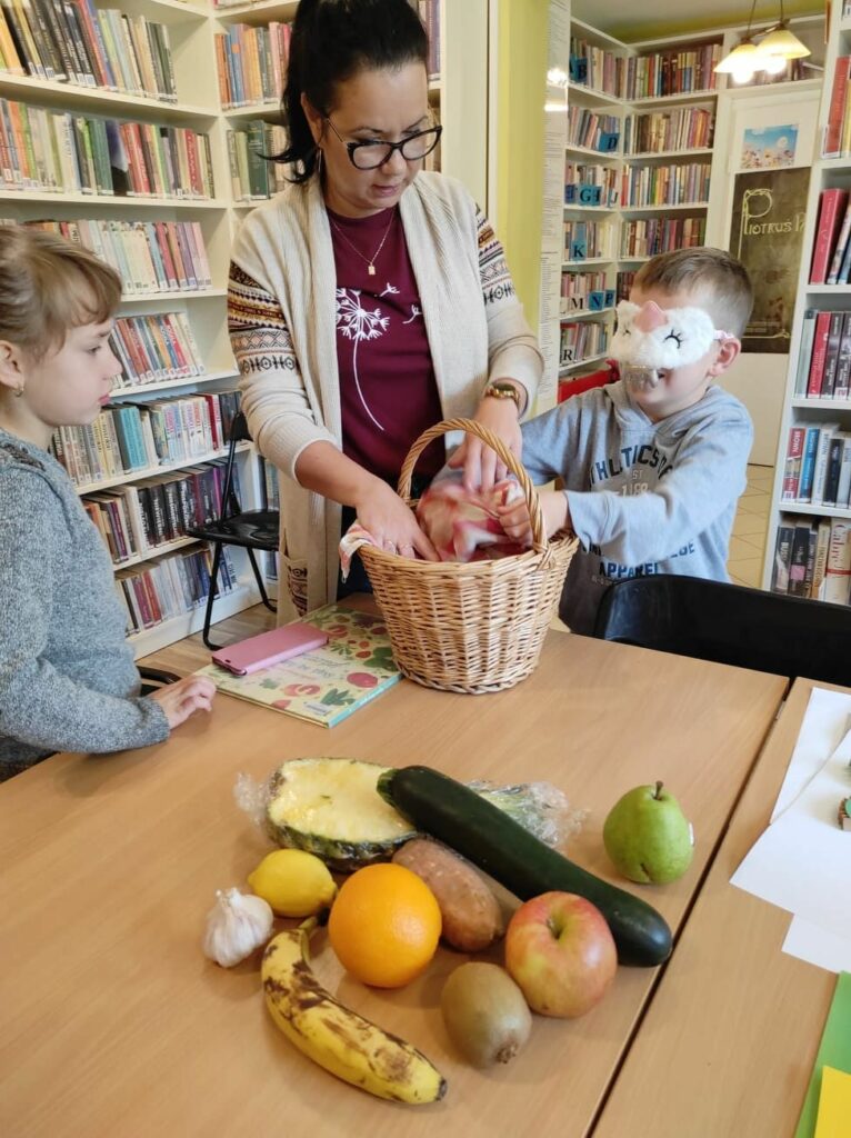 na zdjęciu widać dwoje dzieci dzieci: dziewczynkę i chłopca, widać również bibliotekarkę. Chłopiec ma zasłonięte oczy i próbuje odgadnąć jaki owoc trzyma w rękach. Na stole widać kosz oraz różnego rodzaju owoce i warzywa np. banan, jabłko czy cukinia.