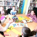 na zdjęciu widać dzieci siedzące wokół stołu oraz bibliotekarkę pokazującą jak wykonać pracę plastyczną. Na stole widać materiały plastyczne, np.: kolorowy papier, nożyczki, kleje, pisaki.