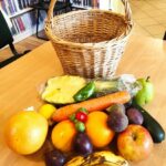 na zdjęciu widać stół na którym stoi wiklinowy kosz a przed nim poukładane są owoce i warzywa takie jak np. ananas, czosnek, cukinia, figi, banan, jabłko itp.